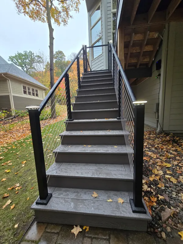 Gray deck stairs with black posts and horizontal cable railings viewed from the yard